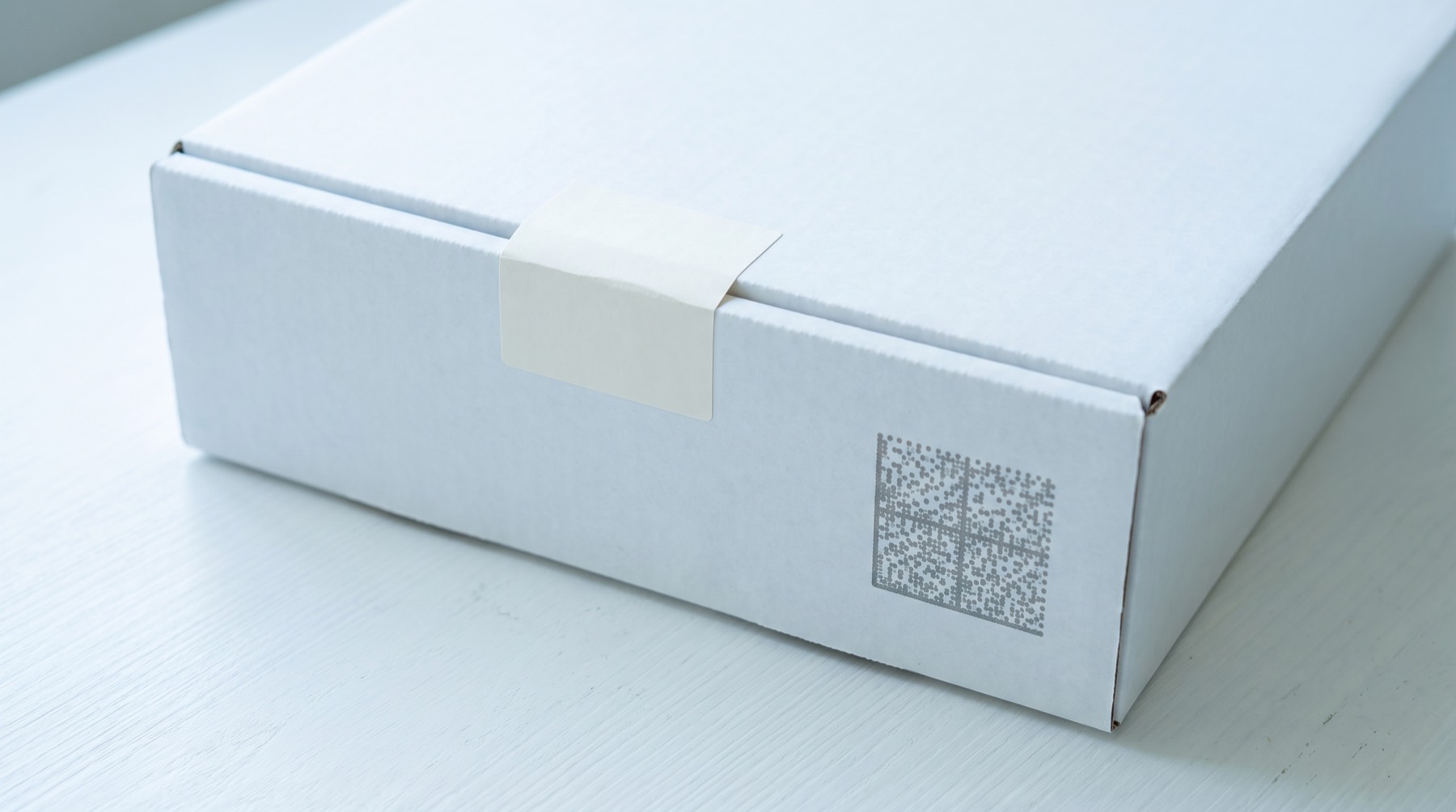 A plain white carton closed with a blank seal strip, a square dot-matrix code printed on its side